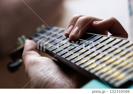 A close-up of a hand playing a ukulele. The hand is positioned on the fretboard, showcasing the strings and frets clearly. The ukulele has a turquoise body.	 132310566