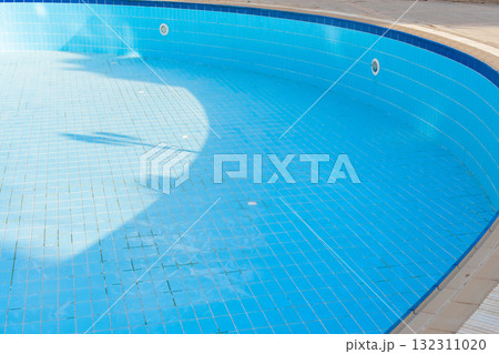 An empty swimming pool with blue tiles and a ladder. The water is absent, revealing the tiled bottom. Surrounding greenery is visible in the background. 132311020