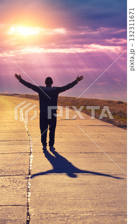 A young man with hands in the air walks along a concrete road and looks at a beautiful orange sunset. Vertical image 132311671