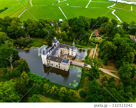 Aerial view of historic castle symmetrical twin towers and central hall framed by forest, moats, and tree lined path, with open fields and residential zone beyond under warm sunrise sky. Aerial view of historic castle symmetrical twin towers and central hall framed by forest, moats, and tree lined path, with open fields and residential zone beyond under warm sunrise sky. 132313242
