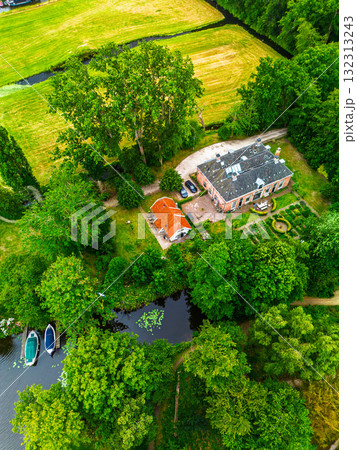 Aerial view of countryside estate brick manor with dark roof and red roofed outbuilding, surrounded by lush greenery, canal with docked boats, gardens and paths in serene rural setting. Aerial view of countryside estate brick manor with dark roof and red roofed outbuilding, surrounded by lush greenery, canal with docked boats, gardens and paths in serene rural setting. 132313243