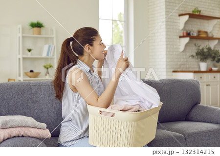 Happy young housewife sitting on sofa with laundry basket and smelling clean freshly washed clothes 132314215