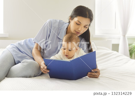 Mother reading a blue board book with baby on a bright bed in warm morning light 132314229