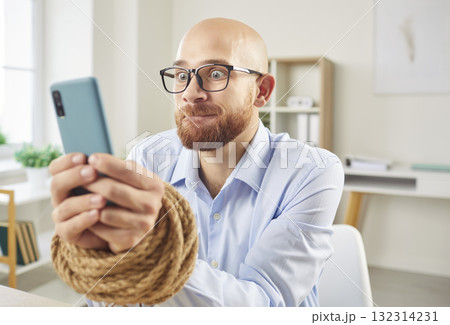 Addicted male employee with tied hands holding mobile phone sitting at the desk in office 132314231