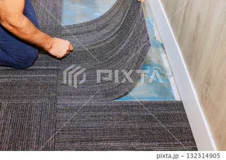 Worker carefully lays down gray carpet tiles in an office on precise alignment installation techniques. Worker carefully lays down gray carpet tiles in an office on precise alignment installation techniques. 132314905
