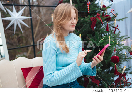Smiling blonde Caucasian woman holding red mug near Christmas tree in cozy festive interior 132315104