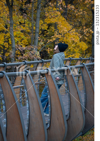 A woman in a green jacket and black hat stands on the ecological trail bridge in VDNH park surrounded by golden autumn leaves A woman in a green jacket and black hat stands on the ecological trail bridge in VDNH park surrounded by golden autumn leaves 132315233