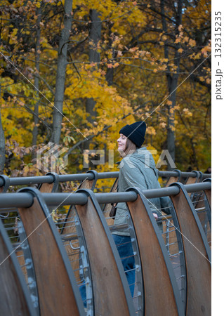 A woman in a green quilted jacket and black hat smiles while standing on a wooden bridge surrounded by yellow autumn foliage in VDNH park 132315235