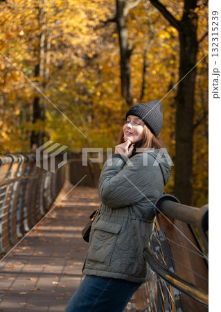 A smiling woman in a green quilted jacket and black hat leans on a wooden bridge surrounded by bright autumn foliage in VDNH park A smiling woman in a green quilted jacket and black hat leans on a wooden bridge surrounded by bright autumn foliage in VDNH park 132315239