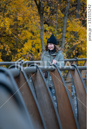 A woman in a green quilted jacket and black hat smiles while standing on a wooden bridge surrounded by yellow autumn foliage in VDNH park A woman in a green quilted jacket and black hat smiles while standing on a wooden bridge surrounded by yellow autumn foliage in VDNH park 132315242