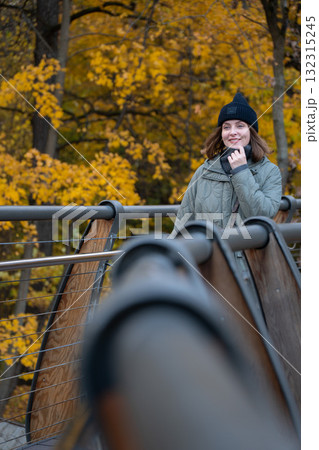 A woman in a green quilted jacket and black hat smiles while standing on a wooden bridge surrounded by yellow autumn foliage in VDNH park 132315245