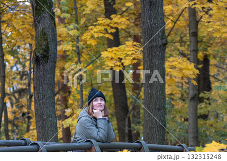 A woman in a green jacket and black hat rests on a wooden bridge railing, surrounded by golden maple leaves and tall trees in VDNH park 132315248