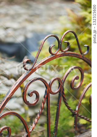 Intricate red metal gate adorned with swirling designs opens to a lush garden surrounded by natural beauty Intricate red metal gate adorned with swirling designs opens to a lush garden surrounded by natural beauty 132315600