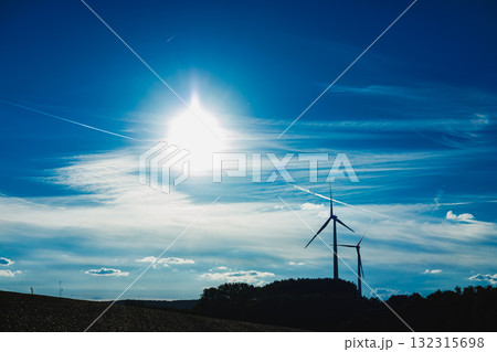 Wind turbines stand tall under a radiant sun in a vibrant sky filled with wispy clouds over a serene landscape during afternoon hours 132315698