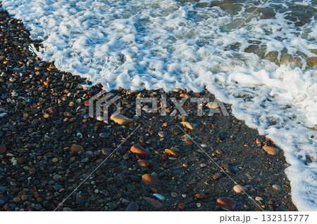 Waves gently rolling onto a colorful pebble beach during a bright sunny day, creating playful foam and a serene atmosphere 132315777