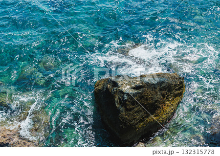 Sunlight glimmers on the clear blue waters, with gentle waves embracing the rugged rocks along the tropical coast at midday 132315778