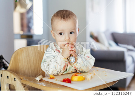 Cute baby learning to eat food with baby spoon by himself on high chair baby led weaning or blw. Infant self-feeding solid food, making mess, fine motor development. Kid enjoying healthy nutrition. Cute baby learning to eat food with baby spoon by himself on high chair baby led weaning or blw. Infant self-feeding solid food, making mess, fine motor development. Kid enjoying healthy nutrition. 132316247