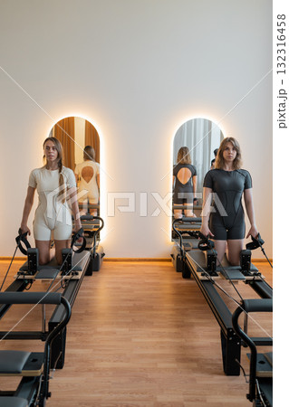 Split training of two girls adjusting reformer arm and core exercises, Woman performing Pilates exercises on reformer machine in gym Split training of two girls adjusting reformer arm and core exercises, Woman performing Pilates exercises on reformer machine in gym 132316458