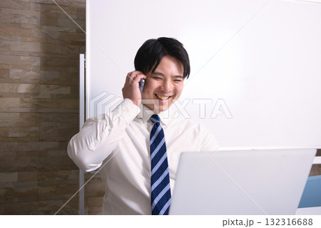 オフィスでパソコンを見ながらスマホで通話する若いビジネスマン 132316688