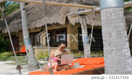 Young woman working on laptop on a tropical beach while her daughter rests nearby Young woman working on laptop on a tropical beach while her daughter rests nearby 132317320