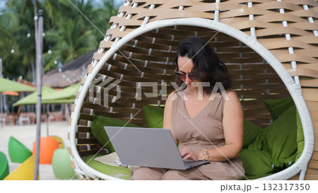 Young woman works on her laptop in a woven chair enjoying a calm tropical beach view embracing the freedom of remote work Young woman works on her laptop in a woven chair enjoying a calm tropical beach view embracing the freedom of remote work 132317350