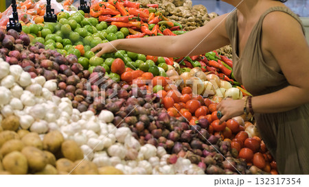 Woman picks green tomatoes from a vibrant display of fresh vegan vegetables in a grocery store focusing on organic healthy meal choices  132317354