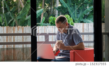 A man enjoys his morning coffee while focused on laptop sitting on a balcony surrounded by greenery in a tropical setting A man enjoys his morning coffee while focused on laptop sitting on a balcony surrounded by greenery in a tropical setting 132317355