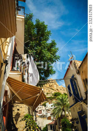 Sunny alley in the Santa Cruz district of Alicante, Spain, with colorful houses, drying laundry, palm trees, and the rocky hill of Santa Barbara Castle in the background. 132317683