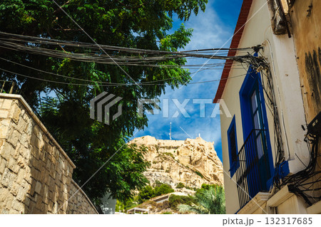 View of the Santa Barbara Castle above the traditional houses of the Santa Cruz district in Alicante, Spain, framed by a stone wall, green trees, and blue sky. 132317685