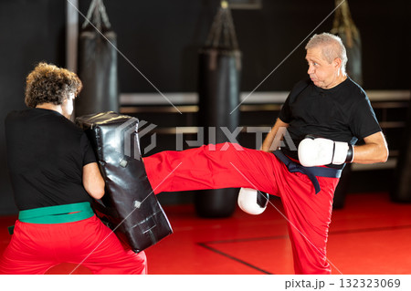 Elderly man practices kicking and punching with a trainer in gym 132323069