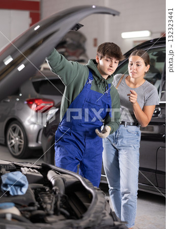Female client talking to young mechanic by open car hood in workshop 132323441