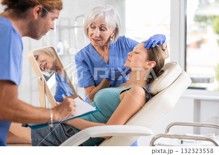 Elderly lady dermatologist examines skin of womans face, male nurse records data in client card 132323558