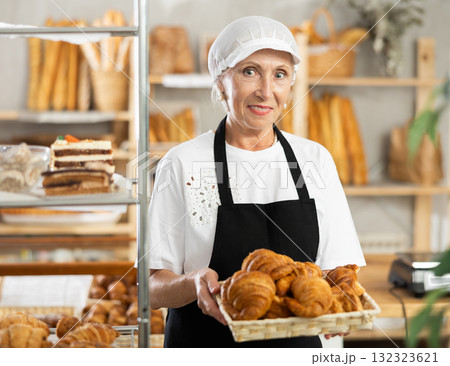 Mature saleswoman displaying croissants in square wicker basket Mature saleswoman displaying croissants in square wicker basket 132323621