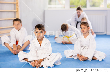 Little children in white kimono sits in a butterfly pose and practices stretching in gym Little children in white kimono sits in a butterfly pose and practices stretching in gym 132323639