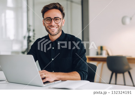 A handsome man in a shirt and glasses is sitting at a white table with a laptop, smiling while working on a digital marketing project in a modern office room with copy space. 132324191
