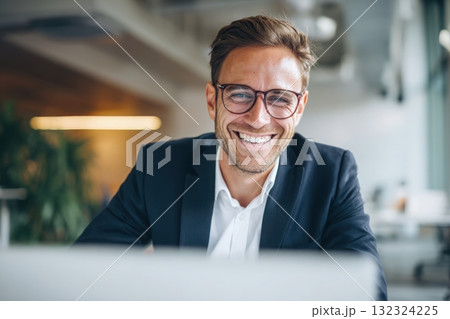 A handsome man in a shirt and glasses is sitting at a white table with a laptop, smiling while working on a digital marketing project in a modern office room with copy space. 132324225