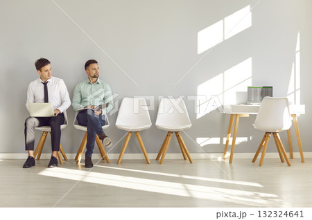 Two men sitting by office wall, waiting for job interview and looking to side at table with laptop 132324641