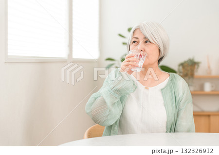 自宅のリビングで水,ミネラルウォーターを飲むグレイヘアーのシニア女性 132326912