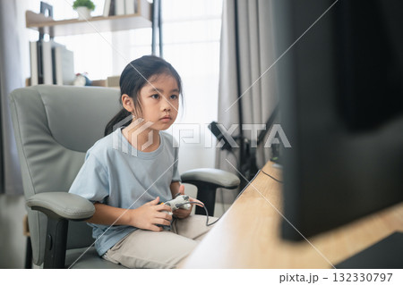 Young girl focused on video game console while sitting in a comfortable chair in a bright room with modern decor and computer setup 132330797