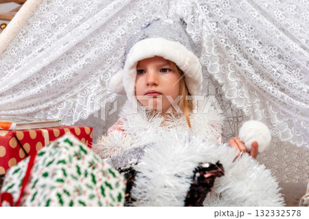 portrait of a cute little girl child wearing a silver color Christmas hat sitting between gift boxes 132332578