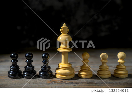 Closeup view of the white chess king standing between white and black chess pawns against a dark background. Concept of leadership or management Closeup view of the white chess king standing between white and black chess pawns against a dark background. Concept of leadership or management 132334011