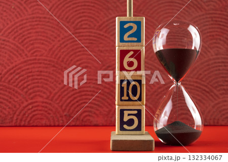 Hourglass with black sand with a wooden number block on a red background. Time passing or time flowing. Sand glass, sand timer, sand clock. Concept of time 132334067