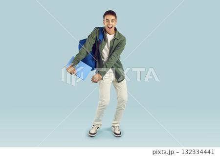 Happy excited student boy holding textbook with backpack isolated on blue studio background. 132334441