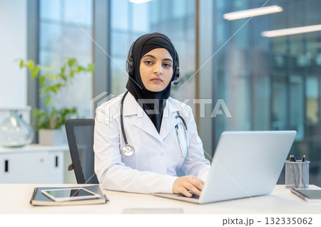 Muslim female doctor wearing a hijab and stethoscope, smiling while sitting at a desk and using a headset and laptop for a remote online medical consultation Muslim female doctor wearing a hijab and stethoscope, smiling while sitting at a desk and using a headset and laptop for a remote online medical consultation 132335062