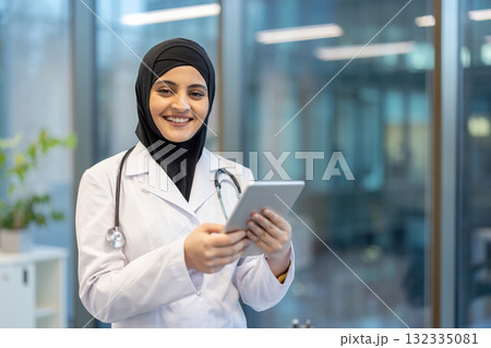 Portrait of a smiling muslim female doctor wearing hijab and lab coat, holding digital tablet and looking at camera, standing in hospital corridor 132335081