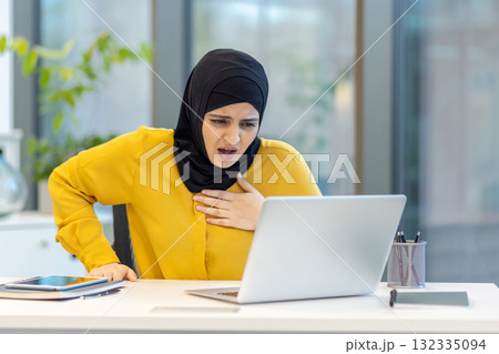 Muslim woman in hijab reacting to shocking online content, experiencing chest pain and stress while sitting at her office desk looking at a laptop computer 132335094