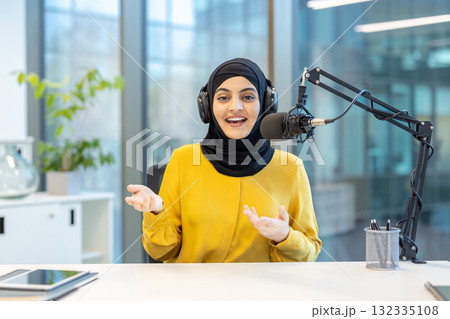 Young muslim woman hosting a radio show, wearing headphones and a hijab, speaking confidently into a microphone while broadcasting live from a modern studio environment 132335108