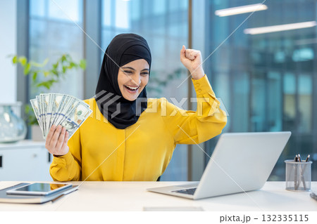 Young muslim woman wearing a hijab celebrates financial achievement, holding cash and looking happily at her laptop while working in a modern office setup Young muslim woman wearing a hijab celebrates financial achievement, holding cash and looking happily at her laptop while working in a modern office setup 132335115