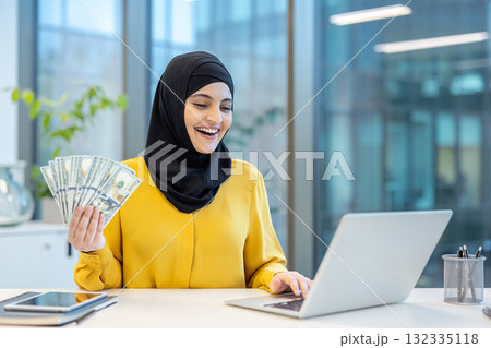 Happy young muslim woman wearing hijab holding us dollar banknotes while using a laptop in a modern office, representing online business success and digital finance 132335118
