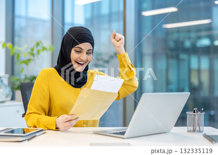 Excited businesswoman wearing hijab is raising her fist while reading good news in a letter at her desk in a modern office, celebrating success and achievement Excited businesswoman wearing hijab is raising her fist while reading good news in a letter at her desk in a modern office, celebrating success and achievement 132335119